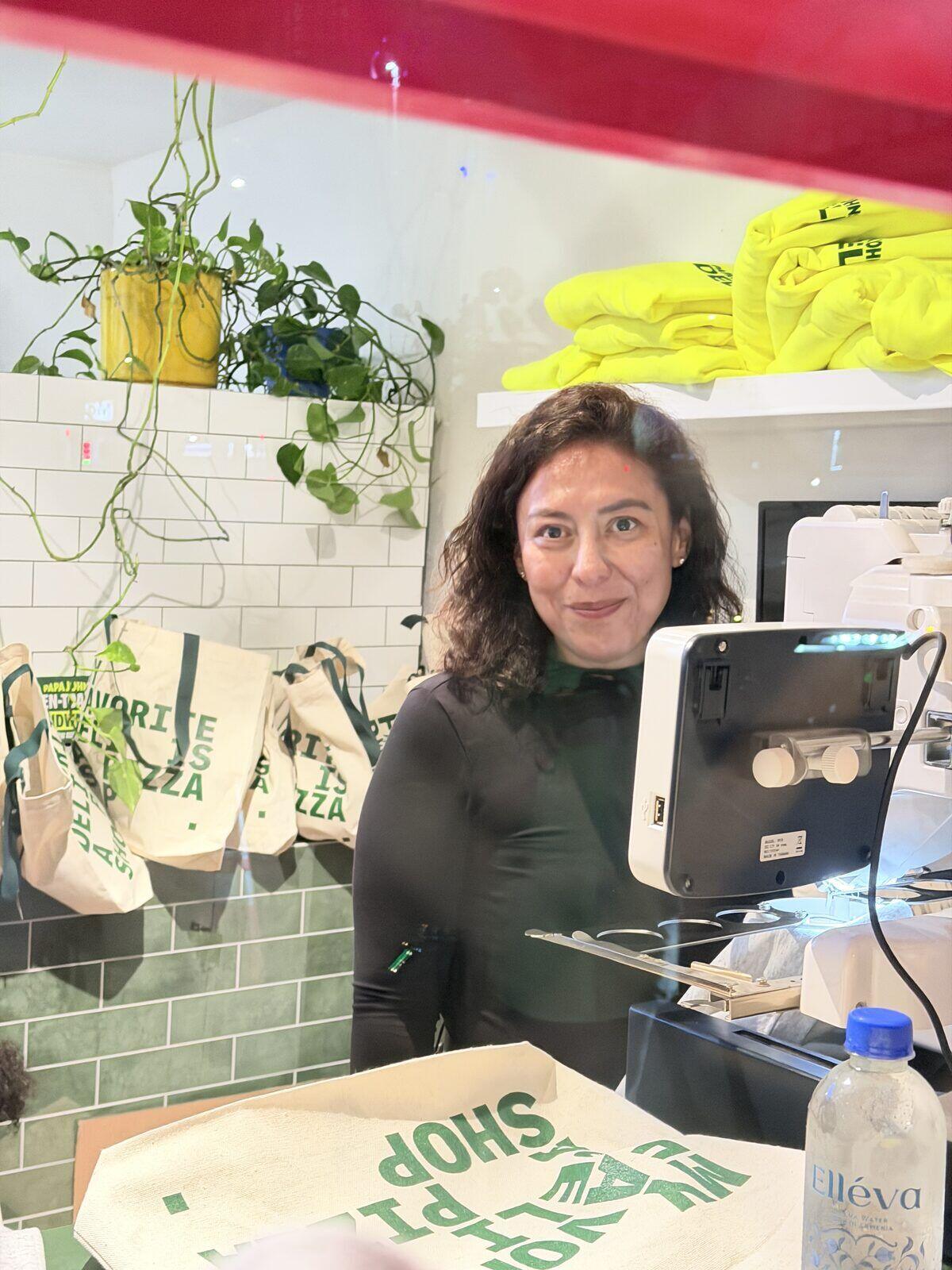 A Merch Troop crew member at a live embroidery machine with stitched totes and neon hoodies behind