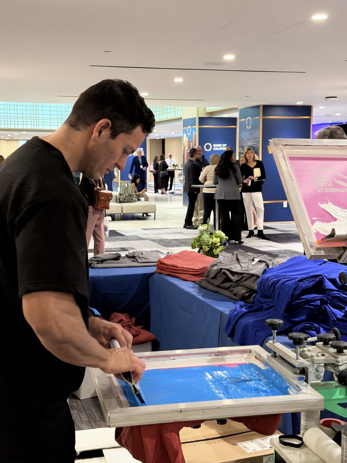 Live screen printing on an LA expo floor — printer mid-pull with guests browsing behind
